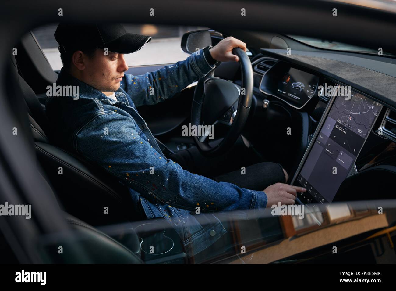 View through open side electric car window. Man in cap sitting in elite automobile, putting one hand on steering wheel and by other hand entering destination address on large touch screen. Stock Photo