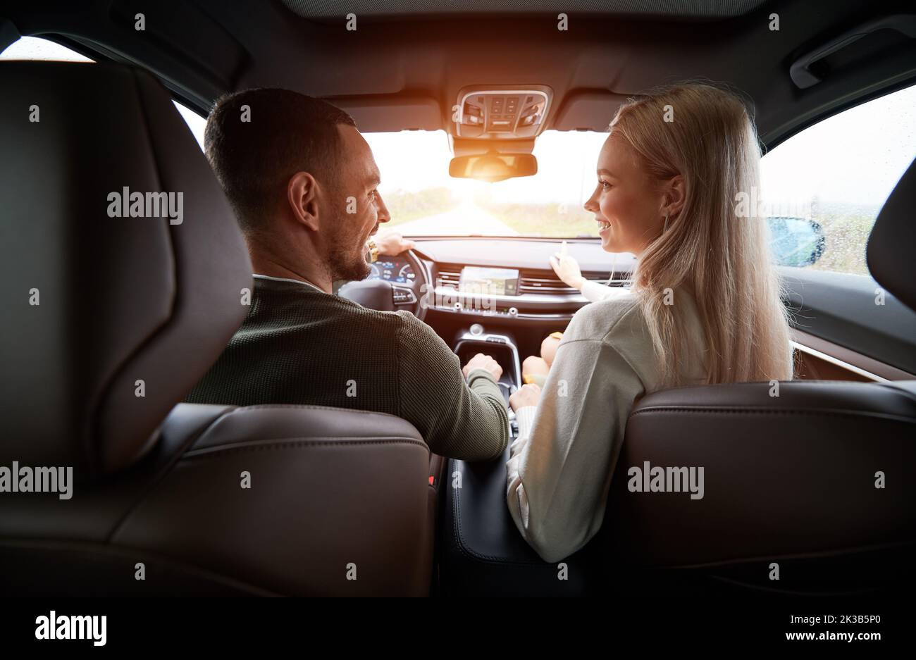 Back view of happy two people inside comfortable car. Joyful driver and cheerful female blonde in the front seat, sincerely smiling and looking at each other. Pleasant atmosphere during auto trip. Stock Photo