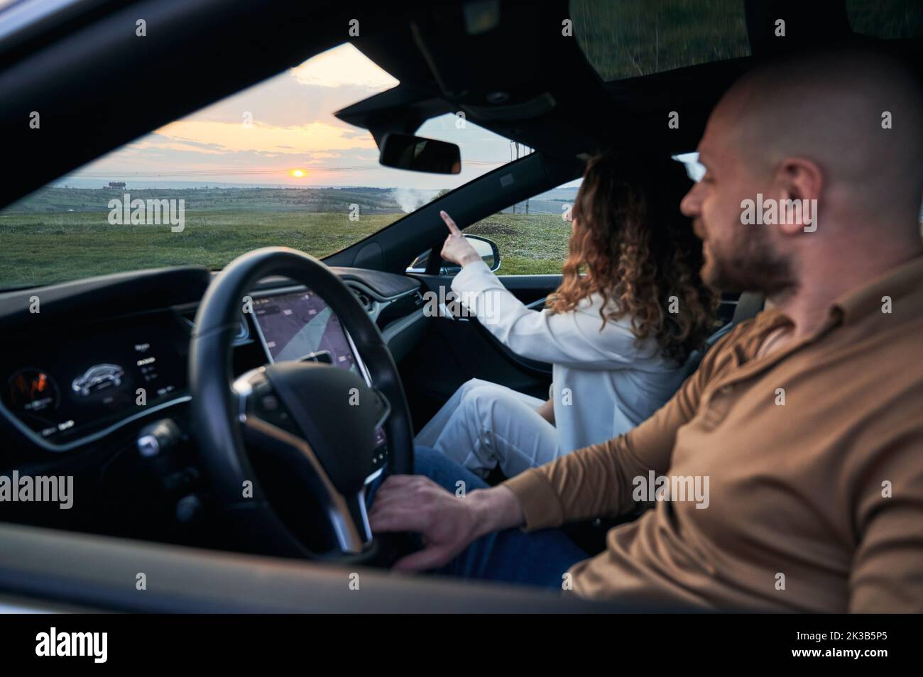 Stylish young woman sitting on passenger seat and pointing finger at sunset while man driving electric car. Female traveler sitting in automobile next to driver and gesturing towards beautiful sky. Stock Photo