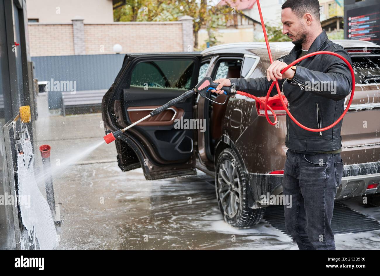 Young man washing car on carwash station outdoor. Handsome worker cleaning automobile, using high pressure soap. Stock Photo