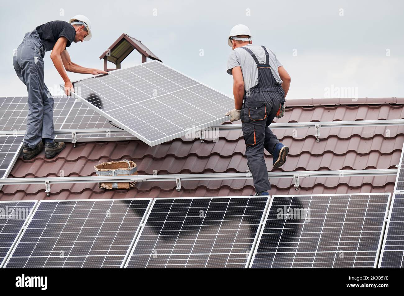 Men technicians lifting up photovoltaic solar moduls on roof of house. Workmen in helmets installing solar panel system outdoors. Concept of alternative and renewable energy. Stock Photo