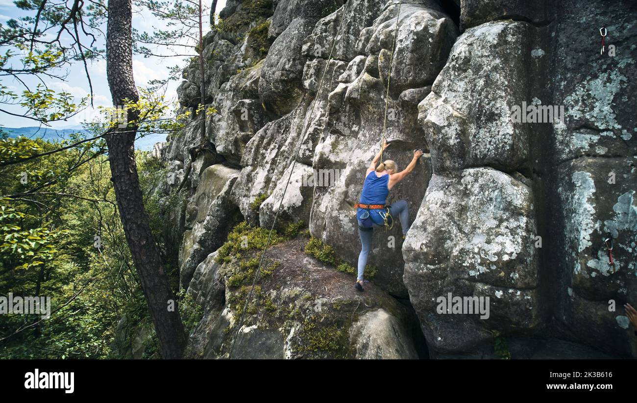 Senior alult woman climbing difficult route on a high rock with rope. Fearless climber training rock climbing on summer day. Concept of extreme sport, adventures and active lifestyle. Aerial view. Stock Photo