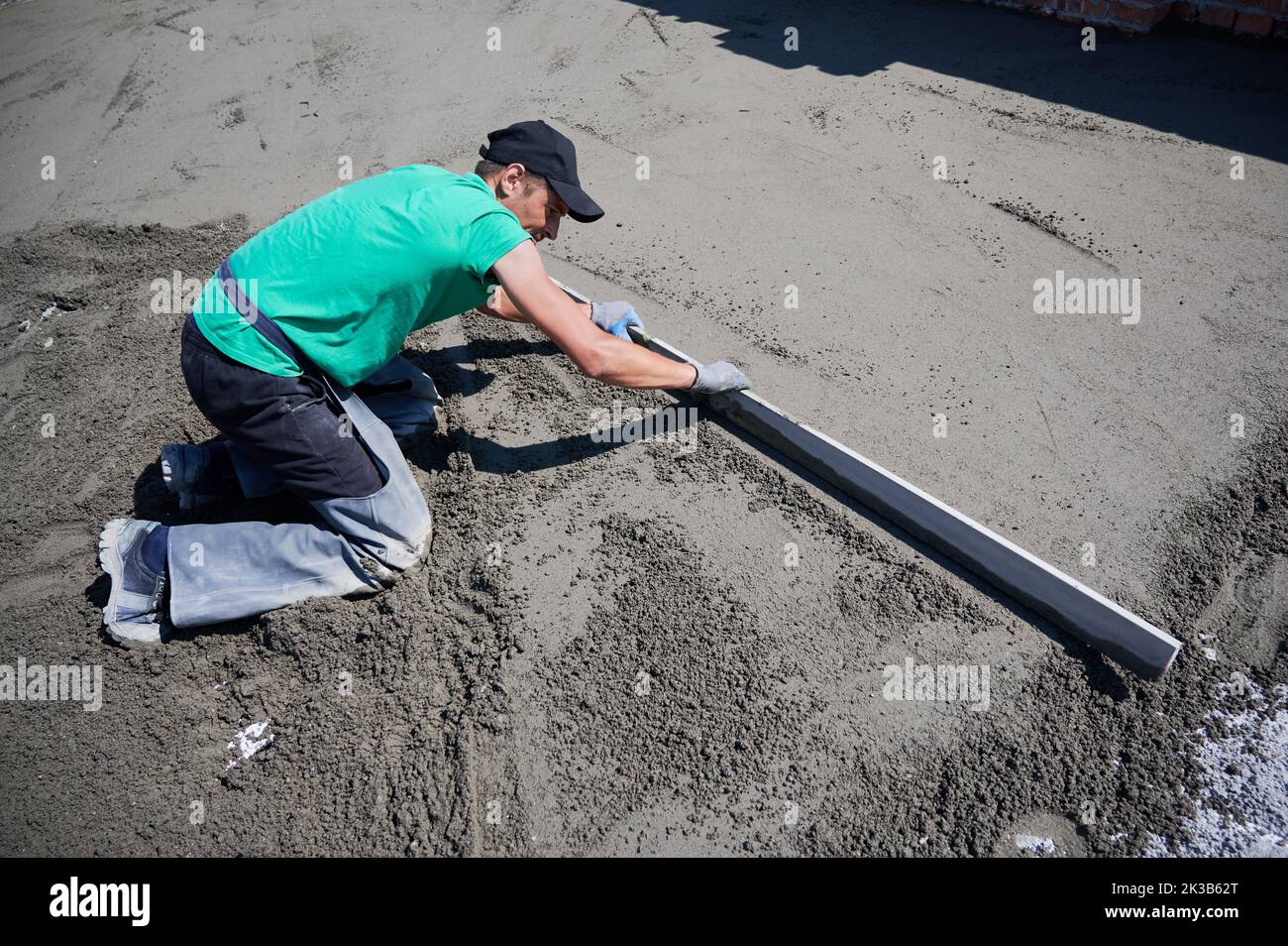 Male worker placing screed rail on the floor covered with sand-cement mix at construction site. Man leveling surface with straight edge while screeding floor outdoors on the roof of new building. Stock Photo