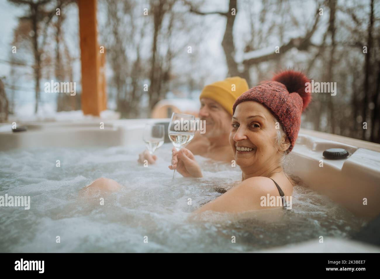Senior couple in kintted cap enjoying together outdoor bathtub and enjoying glass of wine at their terrace during cold winter day. Stock Photo