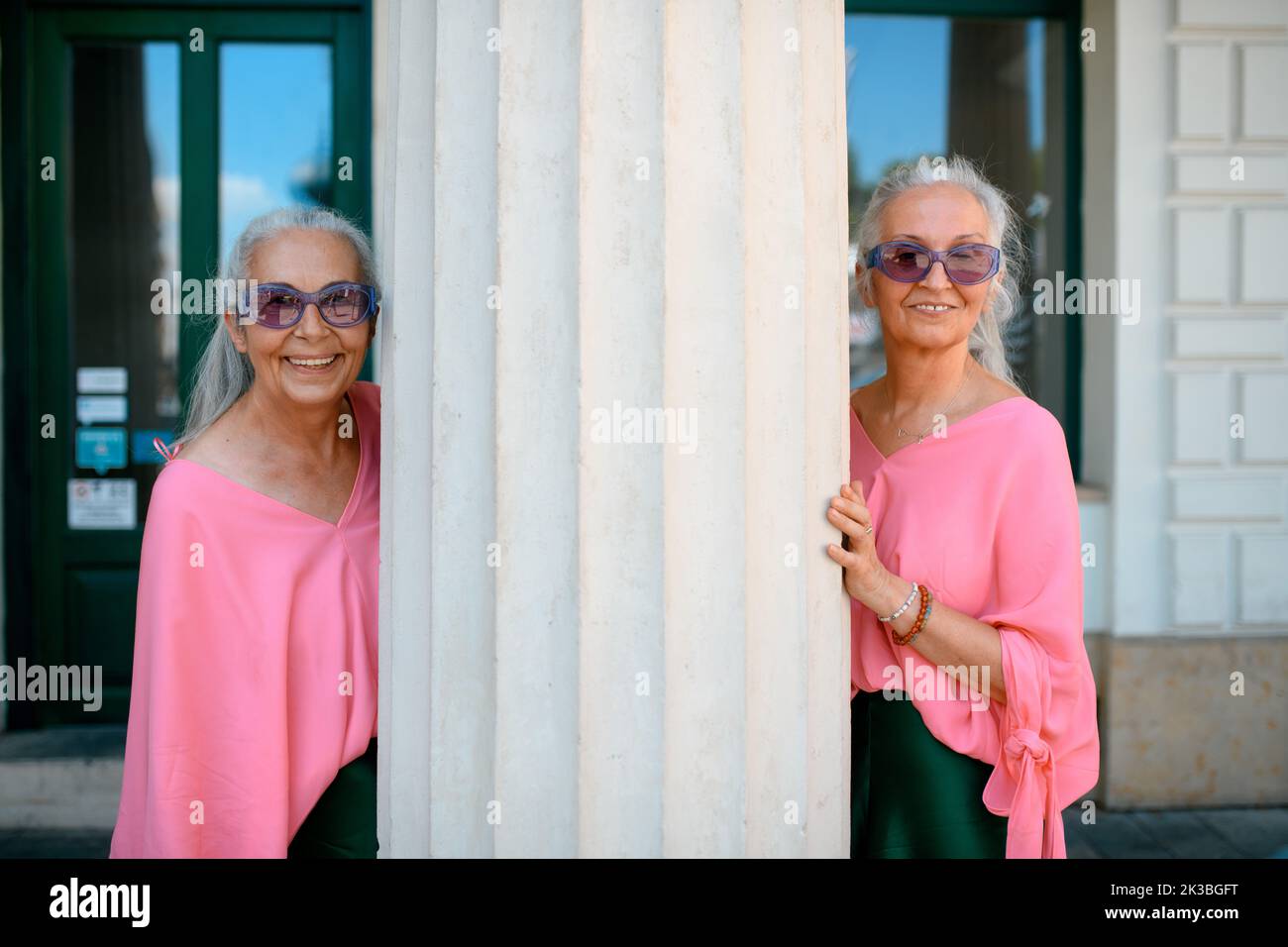 Senior women twins in colourful clothes in city, posing next to architectural column. Stock Photo