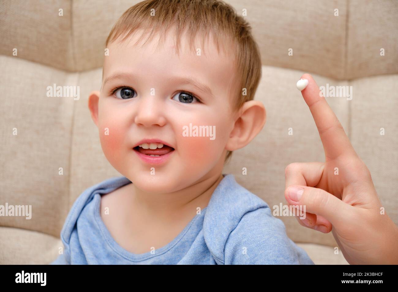 Mother smears allergy cream on face of toddler baby, sofa background in