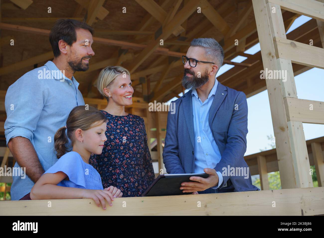 Sales agent showing plans of new unfinished ecologic wooden house to young family on construction site, and looking at view together. Stock Photo