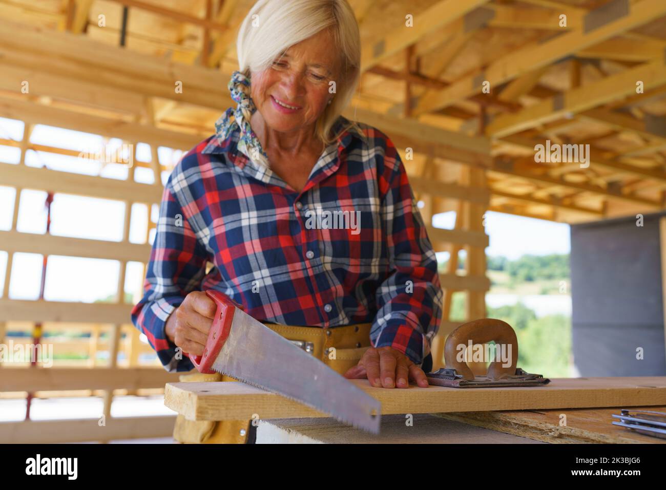 Senior woman sawing wood board inside of her unfinished ecological sustainable wooden eco house. Concept of active independent women and seniors, eco Stock Photo