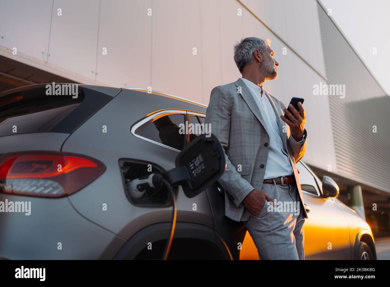 Businessman holding smartphone while charging car at electric vehicle charging station, closeup. Stock Photo