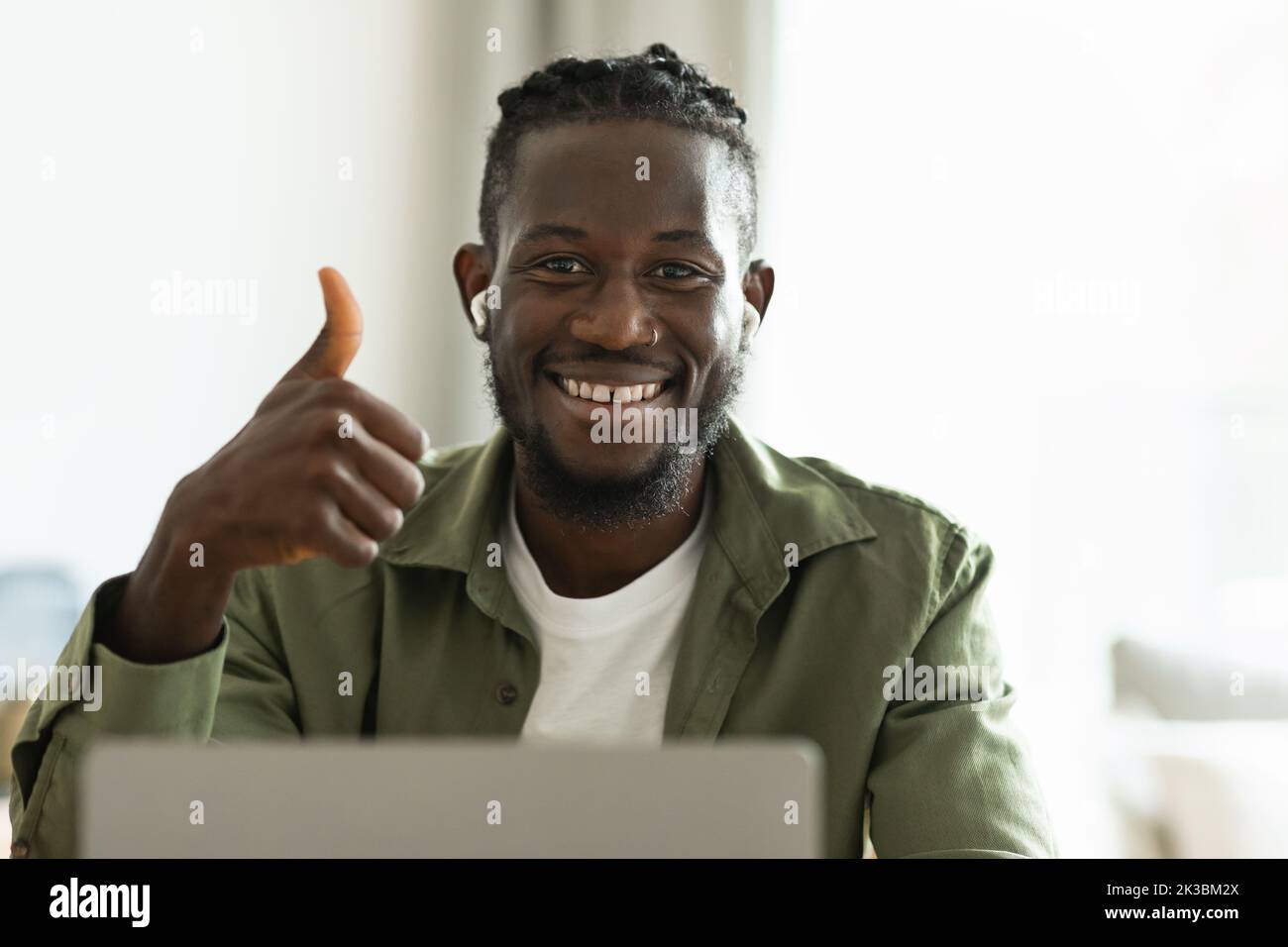 Portrait of positive african american male freelancer working on laptop and gesturing thumbs up, sitting at home office Stock Photo