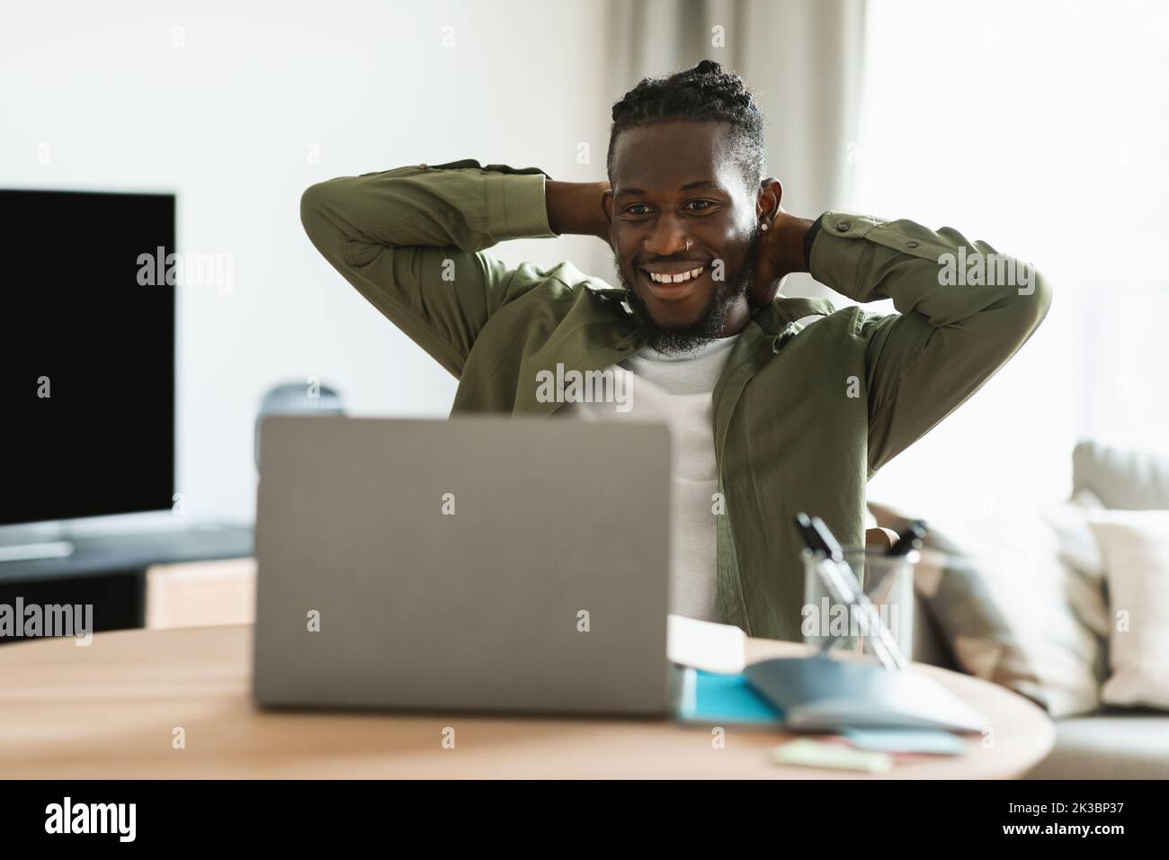Happy black male freelancer looking at laptop computer and relaxing, holding hands behind head, sitting at desk at home Stock Photo