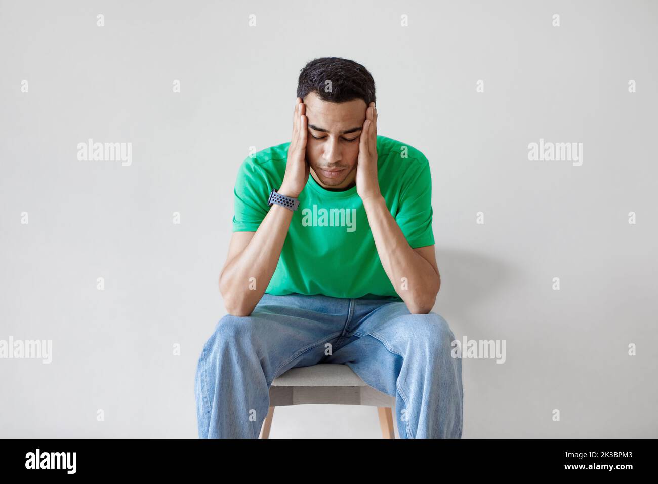 Concerned arab man thinking and worrying about problems, touching head while sitting on chair over light wall Stock Photo