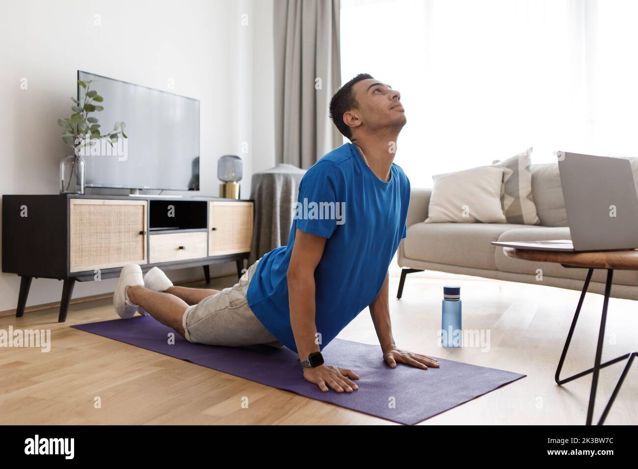 Young arab man practicing pilates with pc, watching online tutorials and standing in cobra pose Stock Photo