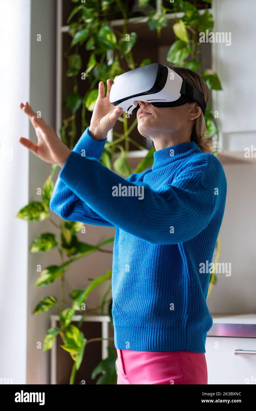 Young woman using VR goggles in cozy interior with plants, hand control, futuristic technologies Stock Photo