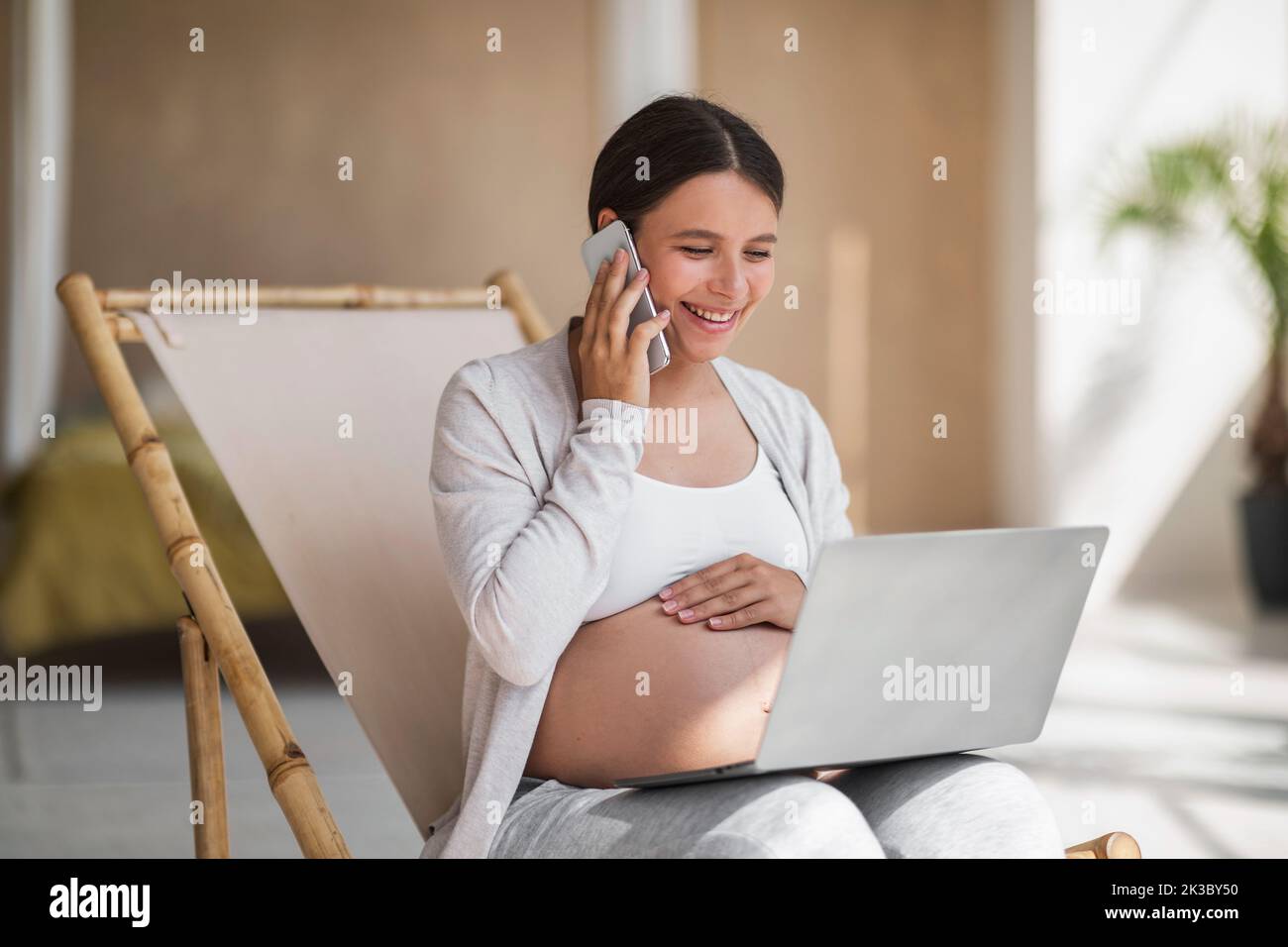 Remote Work Concept. Happy Pregnant Lady Using Laptop And Talking On Cellphone Stock Photo