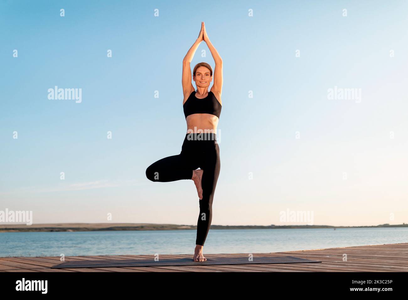 Beautiful middle aged woman standing in tree pose while doing yoga outdoors Stock Photo