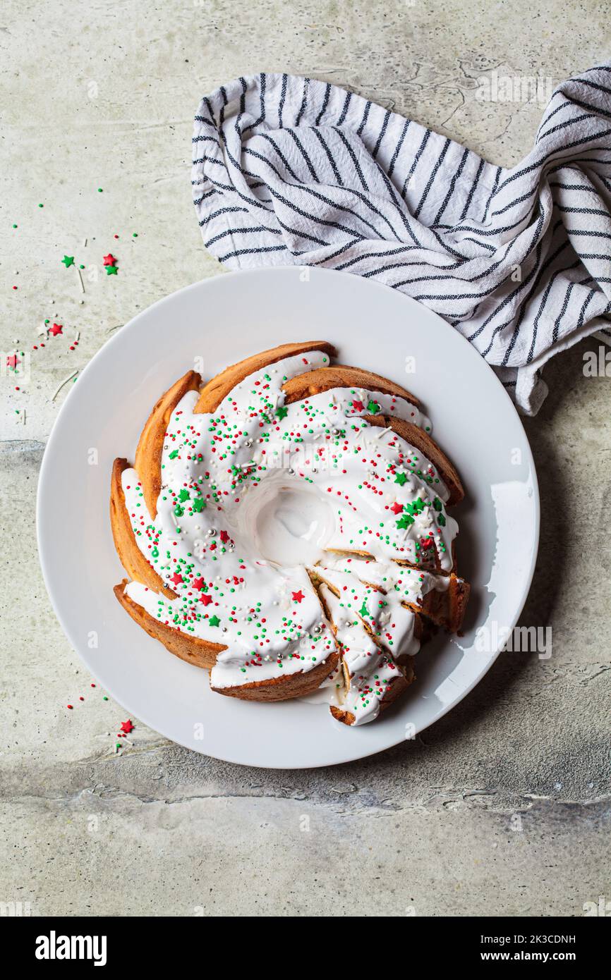 Christmas dessert. Vanilla pound cake with sugar icing and festive colored sprinkles on a white plate, gray background, top view. Stock Photo