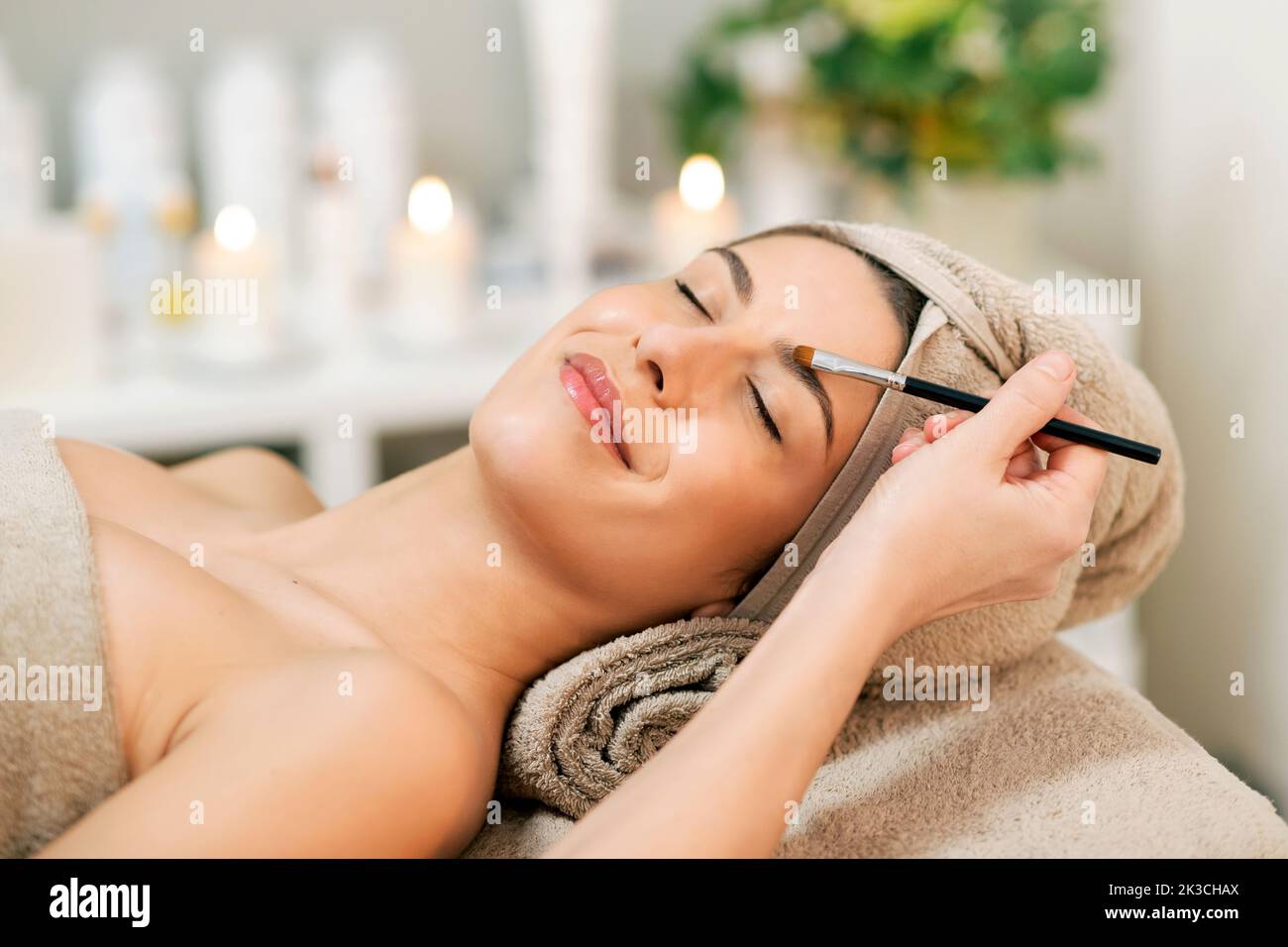 Crop unrecognizable beautician using brush while applying powder on eyebrows of content young female client with perfect skin wrapped in towels and ly Stock Photo