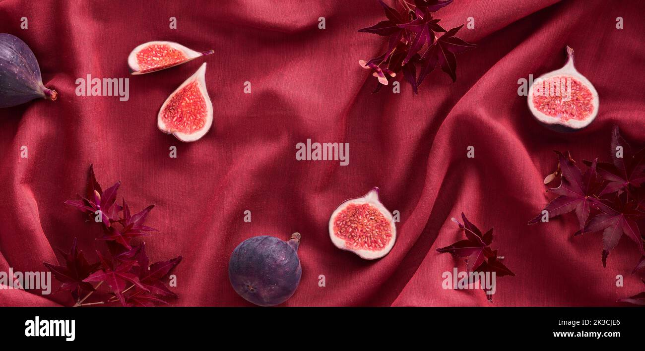 Fresh halved fig fruits and red maple leaves on raspberry red silk textile background. Panoramic banner, seasonal Autumn image. Stock Photo