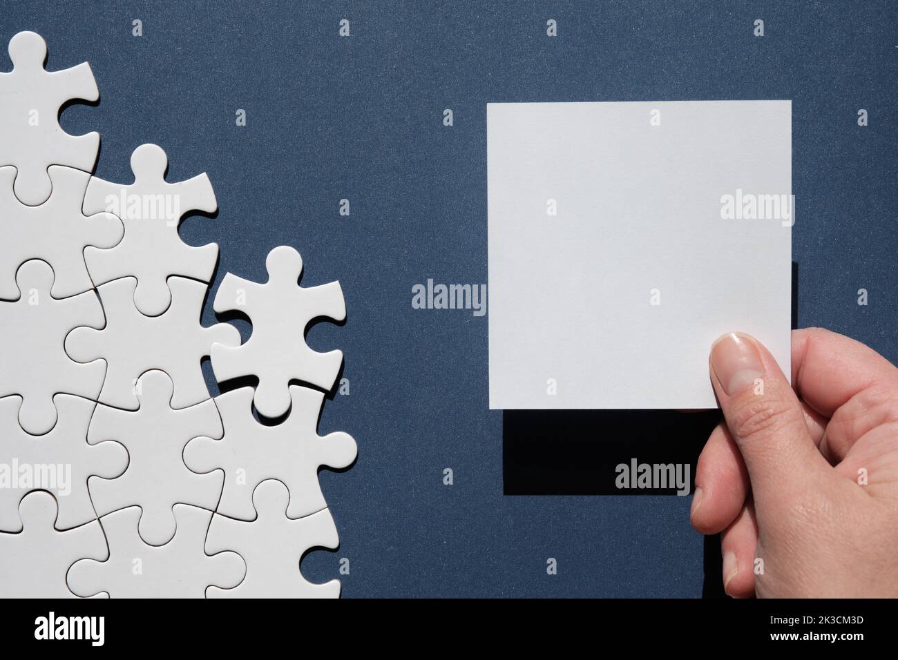Jigsaw puzzle pieces half assembled and separate. Hand hold square blank paper page, space. Top view, flat lay on monochromatic abstract blue paper Stock Photo