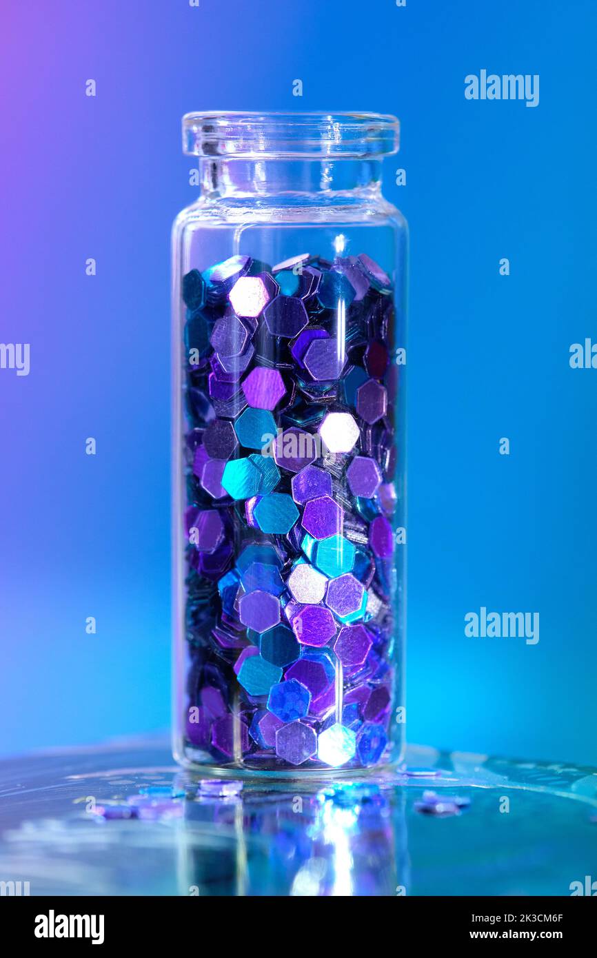Glitter product in transparent bottle on blue background. Hexagone glitter in neon blue, purple and turquoise shades. close-up. Stock Photo