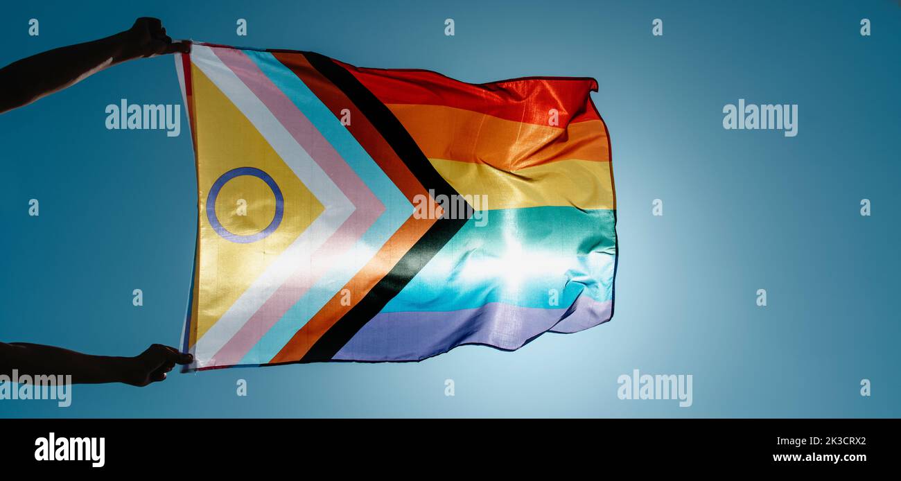 closeup of a man flying an intersex-inclusive progress pride flag on the sky, with the sun in the background, in a panoramic format to use as web bann Stock Photo