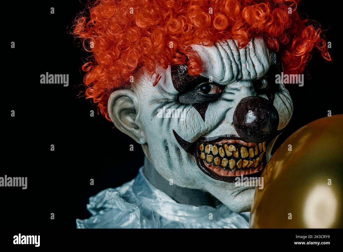 closeup of a creepy evil clown with red hair, staring at the observer, holding a golden balloon in front of him, against a black background Stock Photo