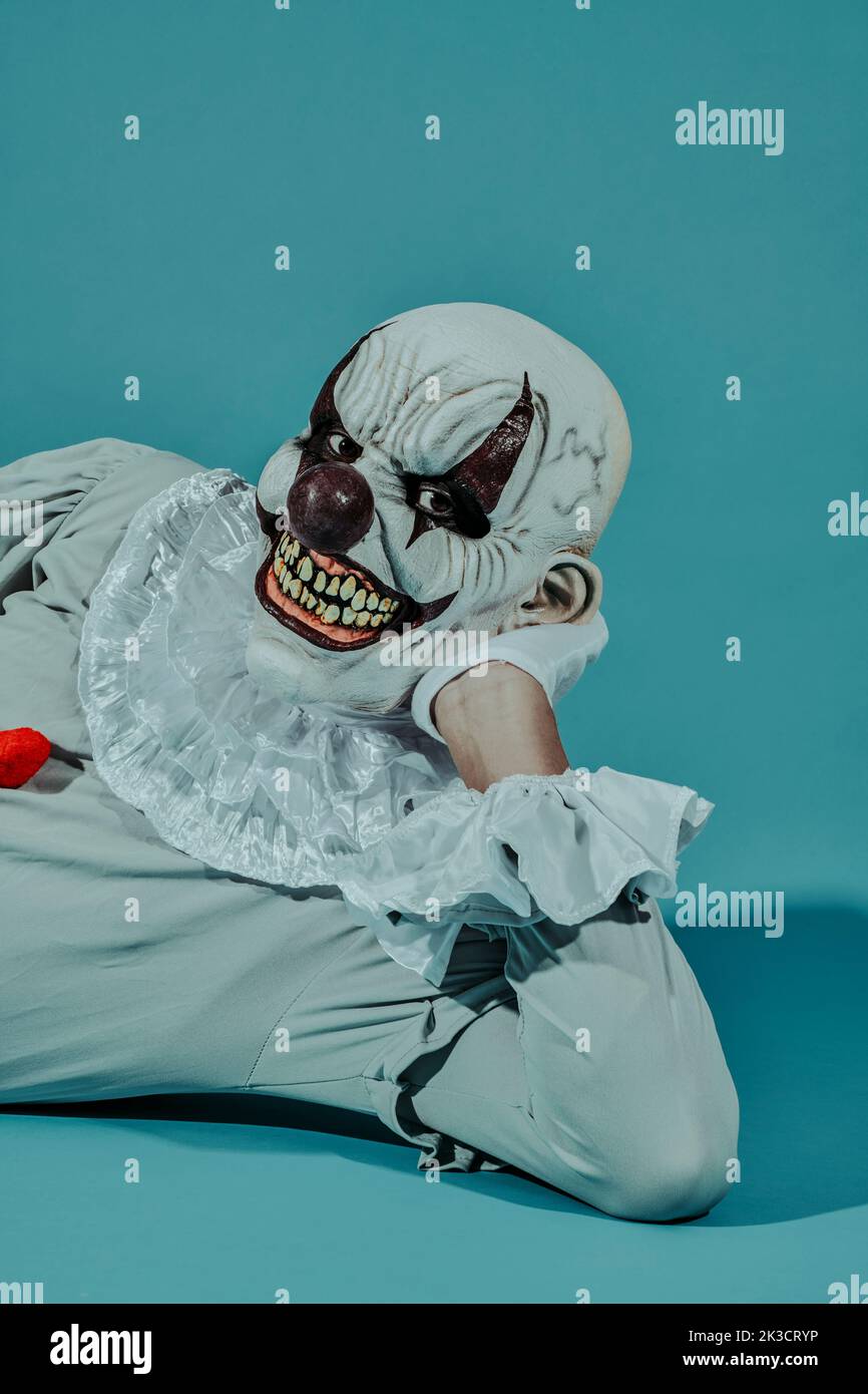 closeup of a creepy bald evil clown, wearing a gray costume with a white ruff, lying on his side, leaning on one arm, on a blue background Stock Photo