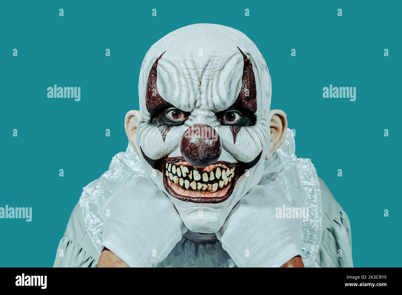 a bald evil clown, wearing white gloves and a costume with a white ruff, staring at the observer with a creepy smile, leaning his head in his hands, o Stock Photo