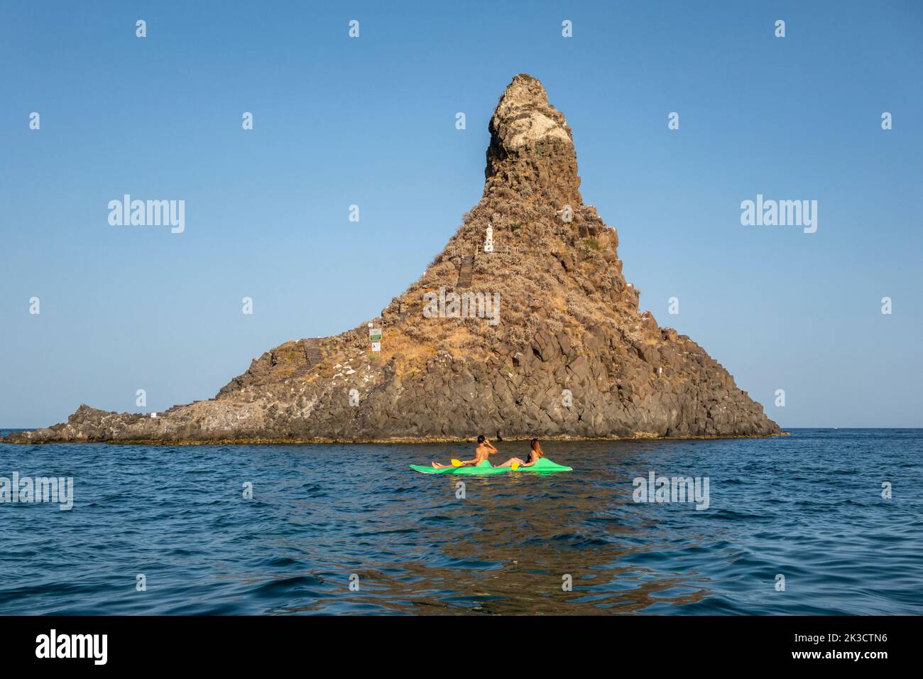 Two kayakers in front of Faraglione Grande, one of the Faraglioni or Isole dei Ciclopi, a group of volcanic basalt sea stacks off Aci Trezza, Sicily Stock Photo