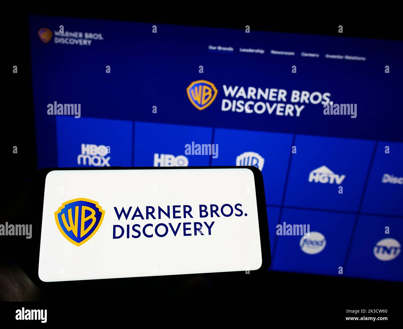 Person holding mobile phone with logo of media company Warner Bros. Discovery Inc. (WBD) on screen in front of web page. Focus on phone display. Stock Photo