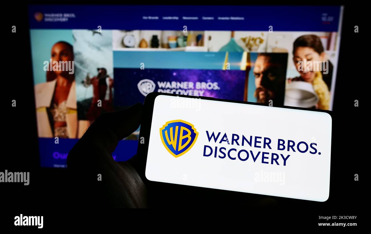 Person holding cellphone with logo of media company Warner Bros. Discovery Inc. (WBD) on screen in front of webpage. Focus on phone display. Stock Photo