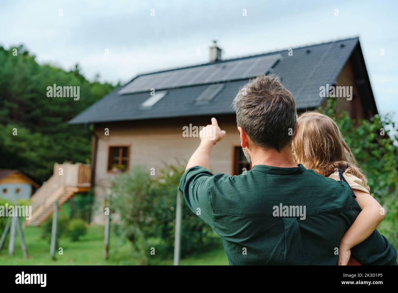 Rear view of dad holding her little girl in arms and showing at their house with solar panels.Alternative energy, saving resources and sustainable lif Stock Photo