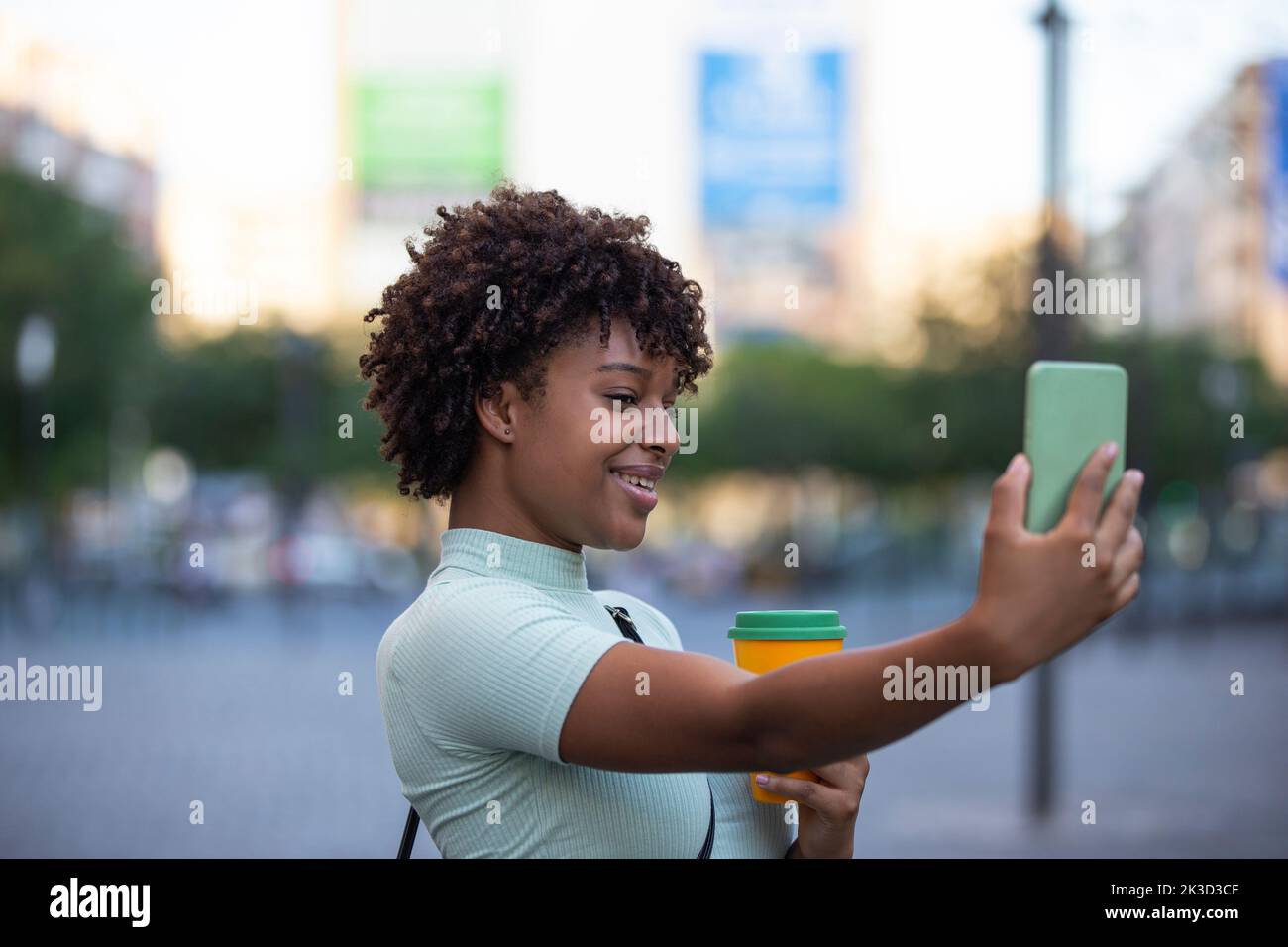 making selfie. Young woman with curly hair wearing fashionable clothes outdoors in the city. Stock Photo