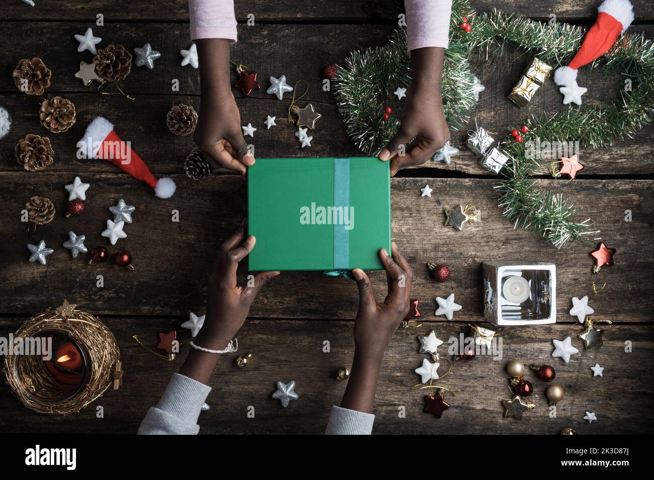 Top view of two coloured kids hands holding a green christmas holiday present box placed on a festive decorated wooden desk with stars, candles and ba Stock Photo