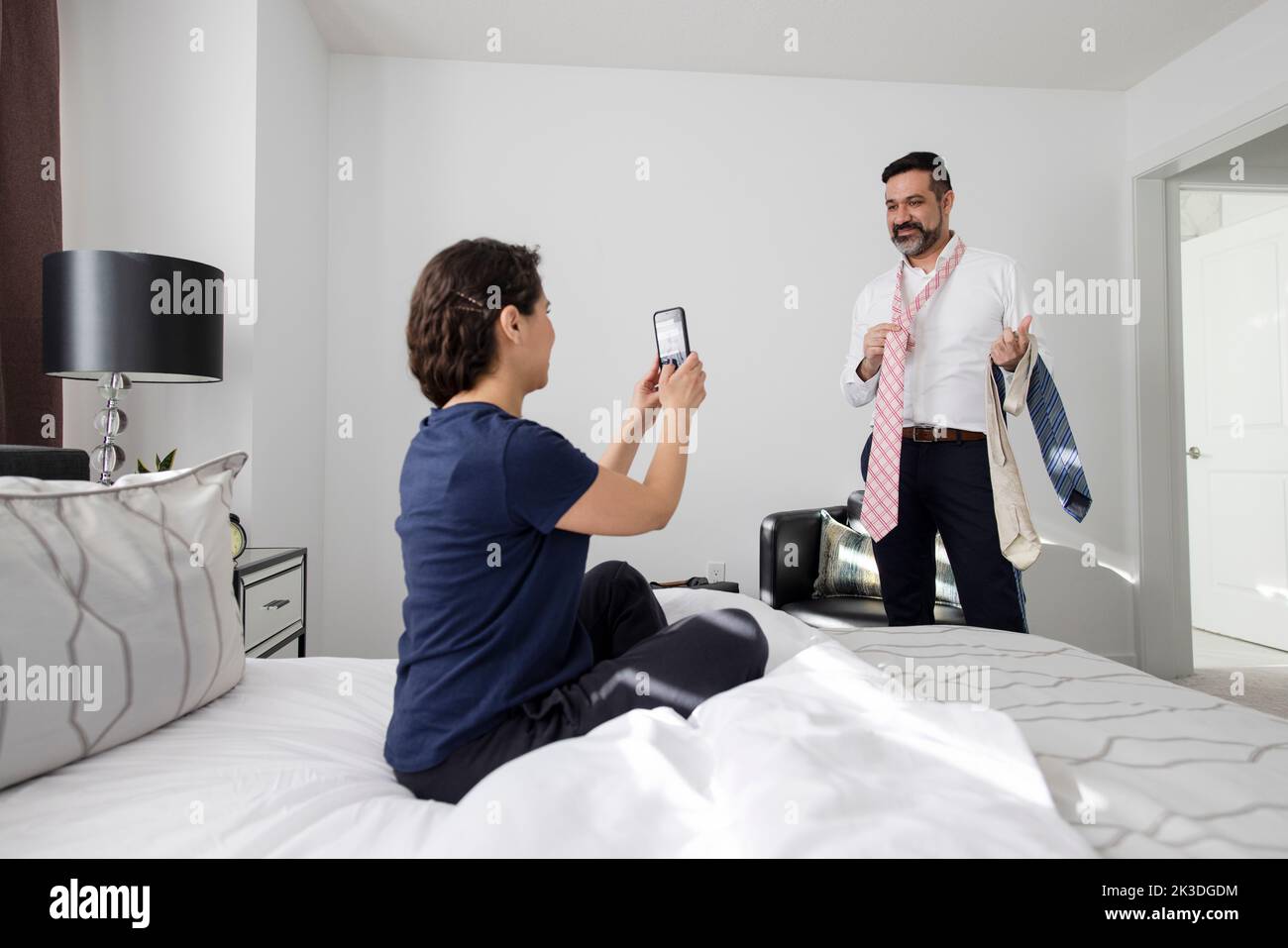 Wife with smart phone photographing husband trying on ties in bedroom Stock Photo