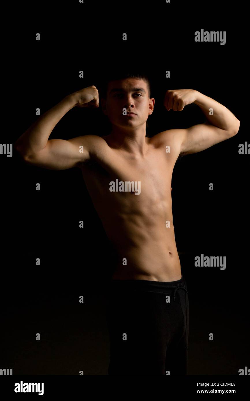 Shirtless 19 year old teenage boy flexing his arms with a black background Stock Photo