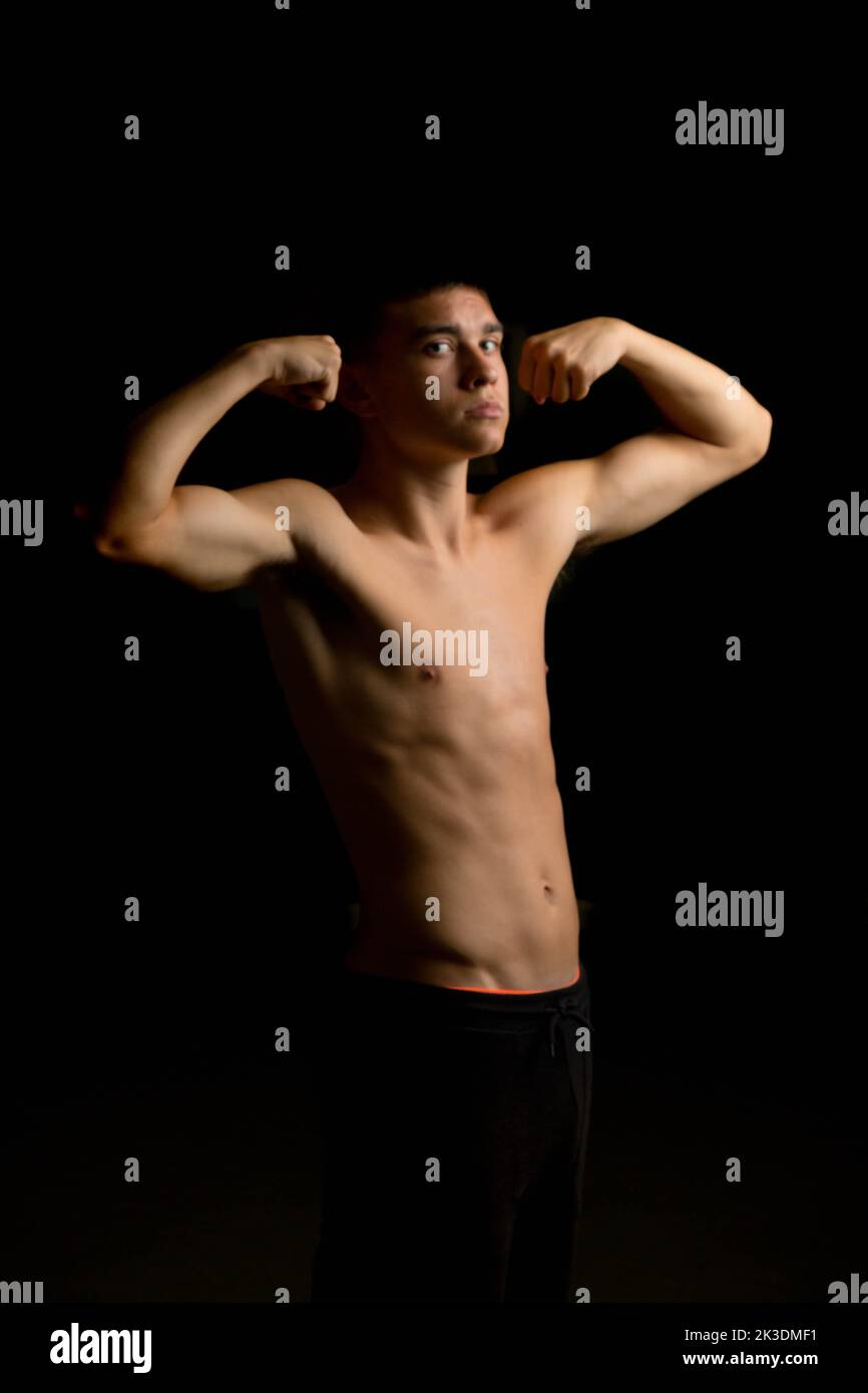 Shirtless 19 year old teenage boy flexing his arms with a black background Stock Photo