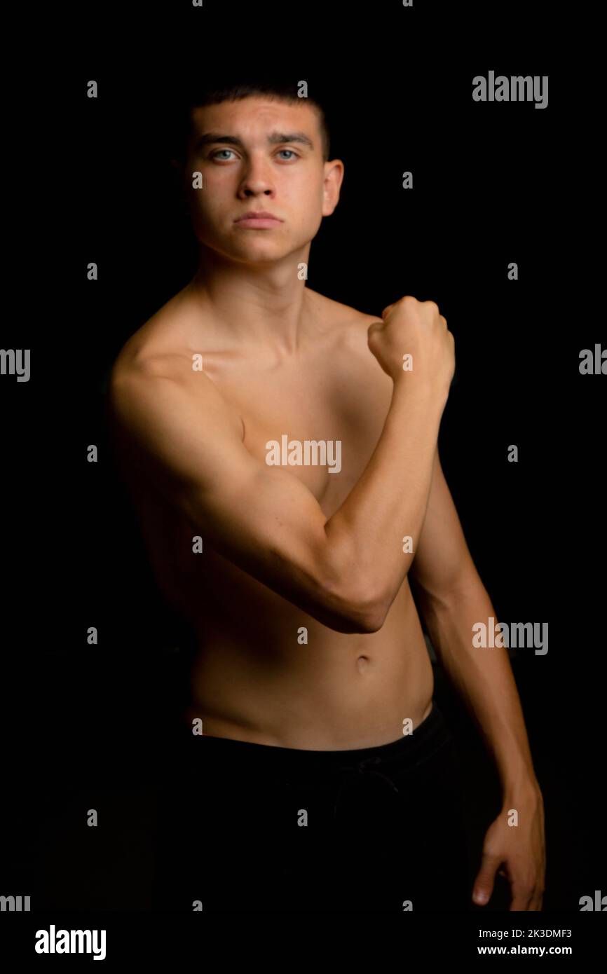Shirtless 19 year old teenage boy flexing his arm muscles on a black background Stock Photo
