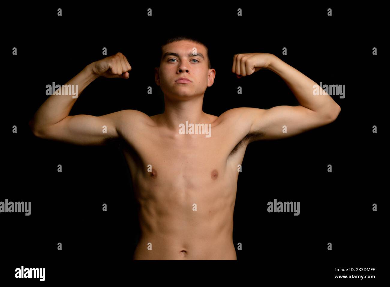 Shirtless 19 year old teenage boy flexing his arm muscles on a black background Stock Photo