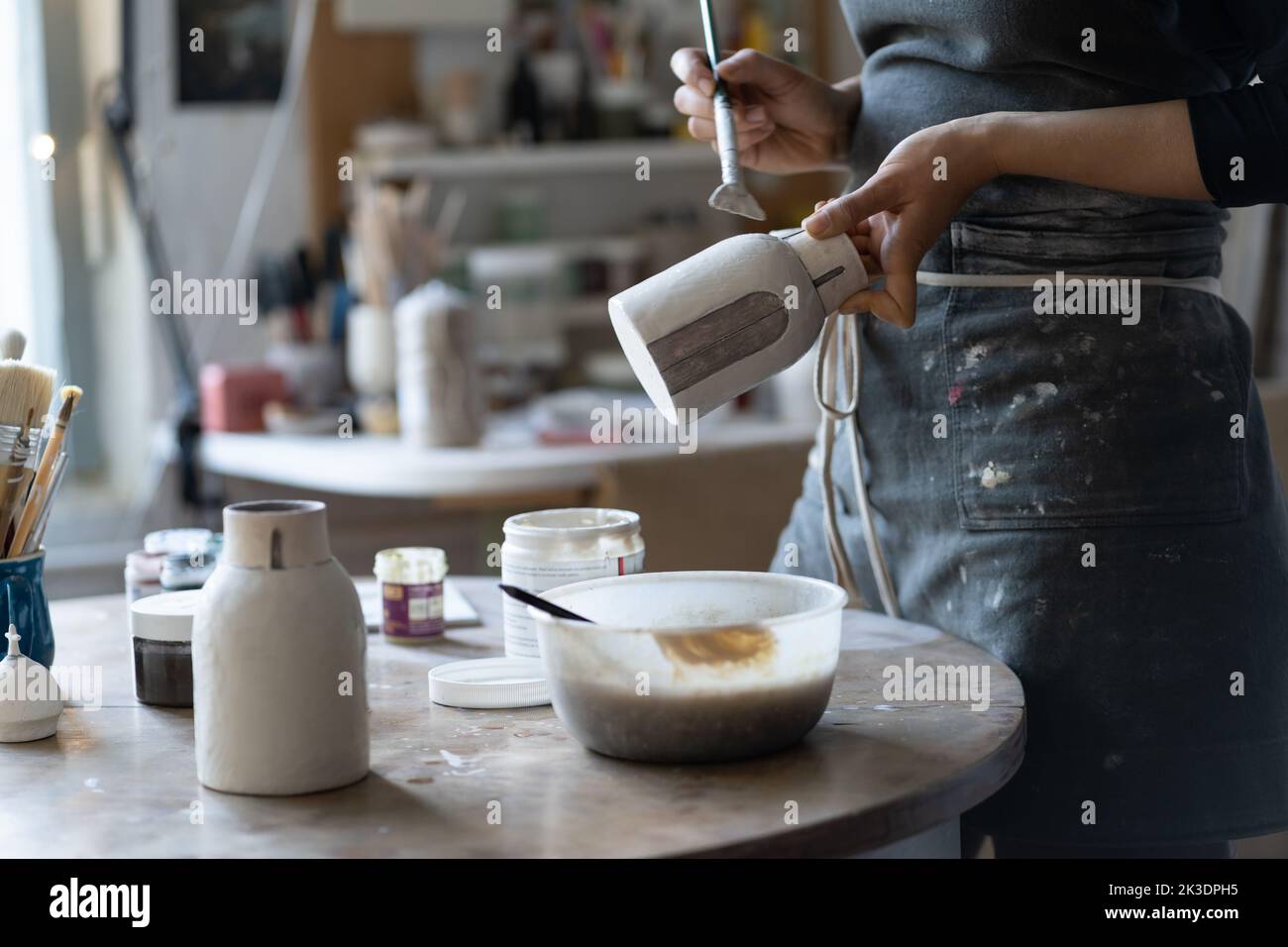 Ceramist in black apron paints clay vase with thick paintbrush near table in cozy workshop Stock Photo