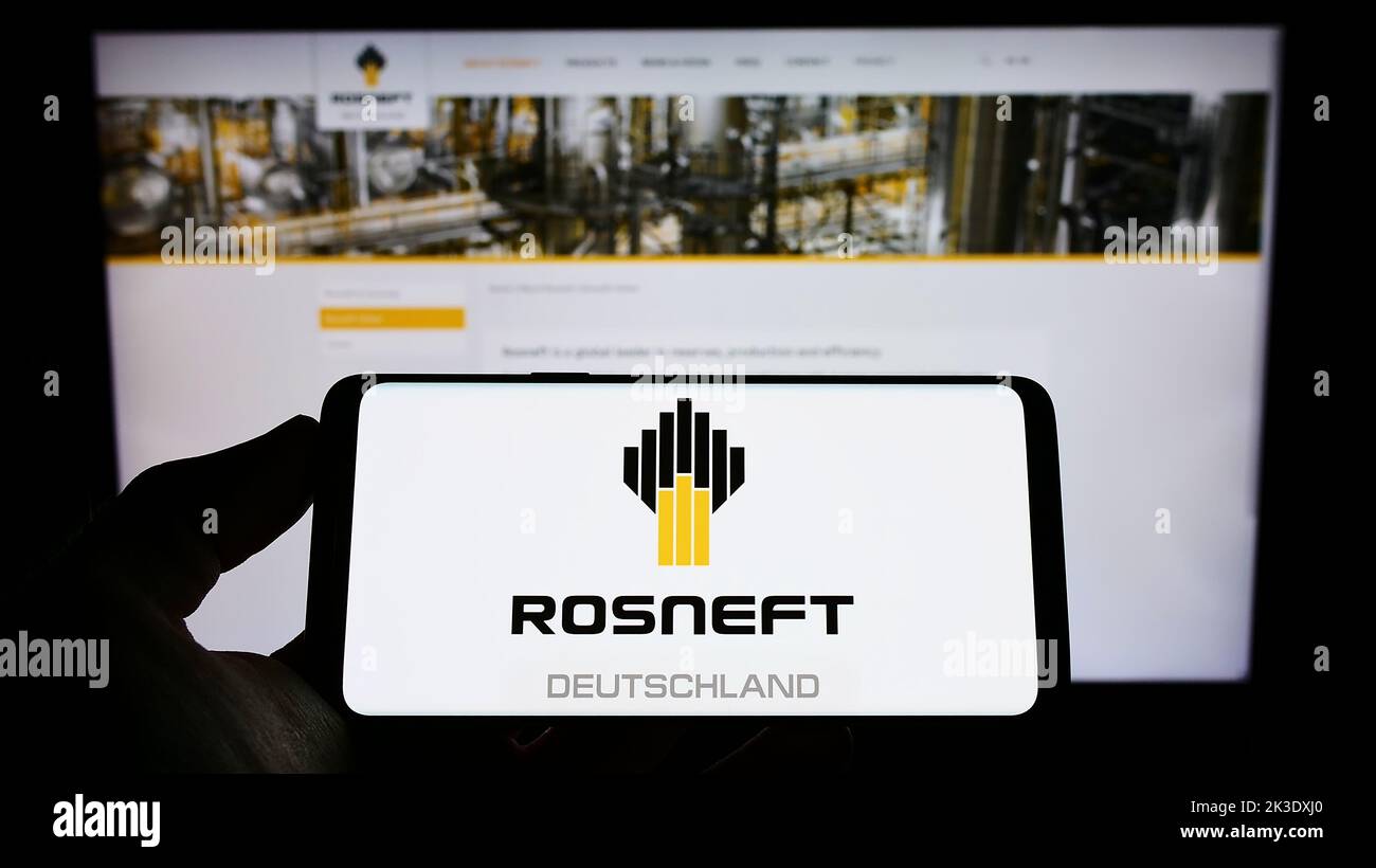 Person holding mobile phone with logo of oil refining company Rosneft Deutschland GmbH on screen in front of web page. Focus on phone display. Stock Photo