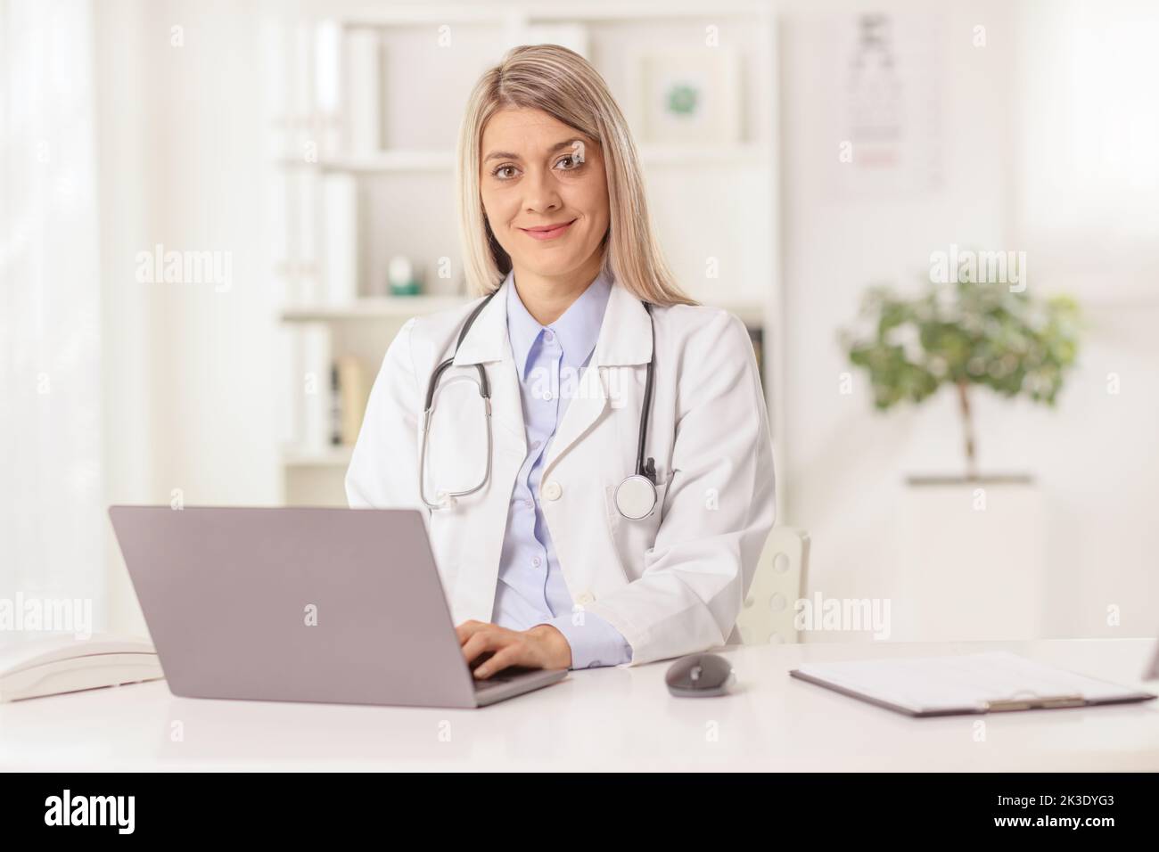 Young female doctor sitting in an office with a laptop computer, typing and smiling at camera Stock Photo