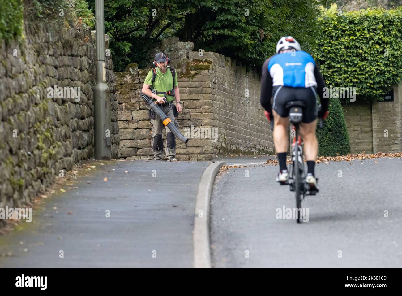 Man using a hand-held leaf blower to blow leaves off the back and out of the way of a cyclist approaching, UK Stock Photo