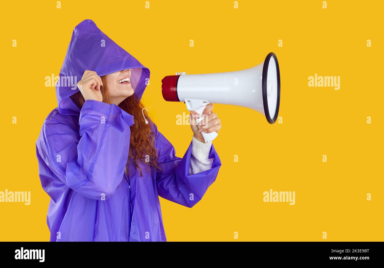 Cheerful joyful woman in waterproof purple raincoat with hood makes announcement using megaphone. Stock Photo