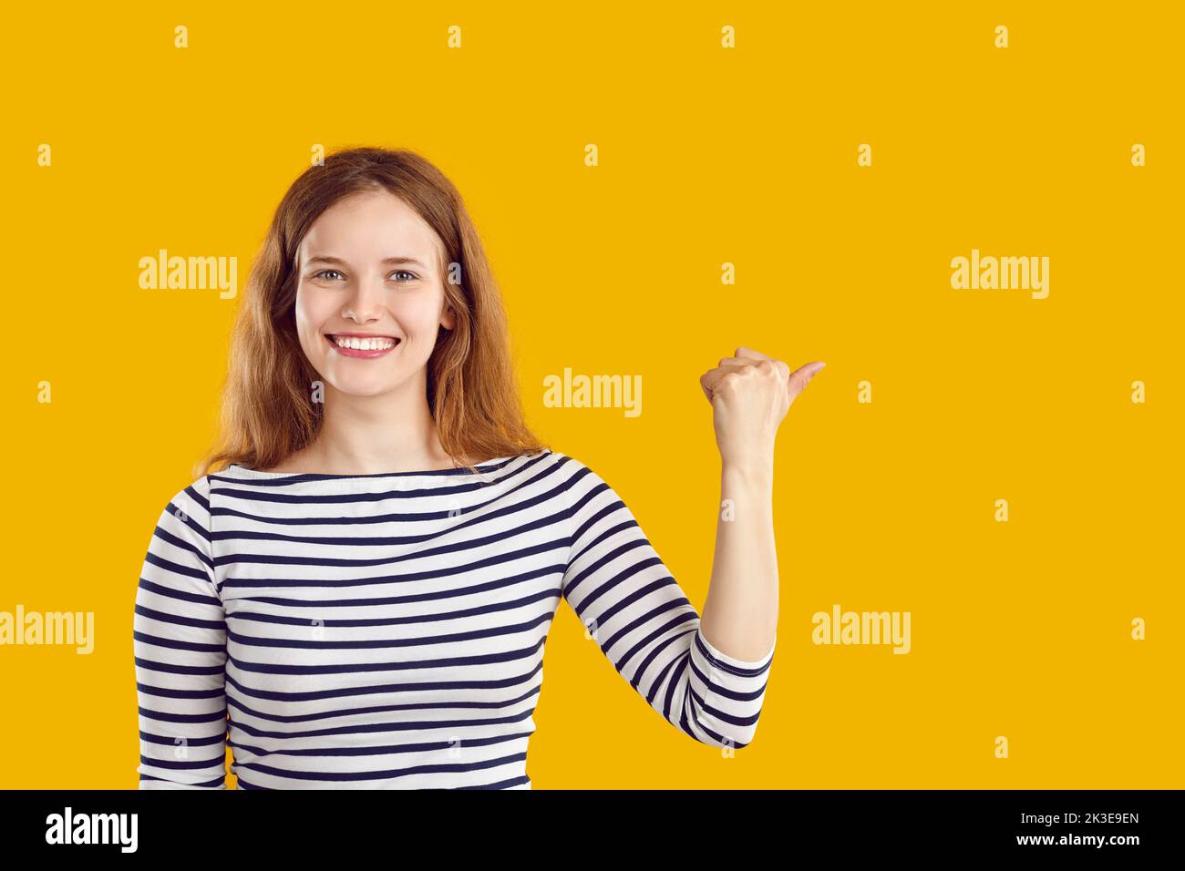 Happy woman smiles and points her thumb to copy space side on yellow studio background Stock Photo