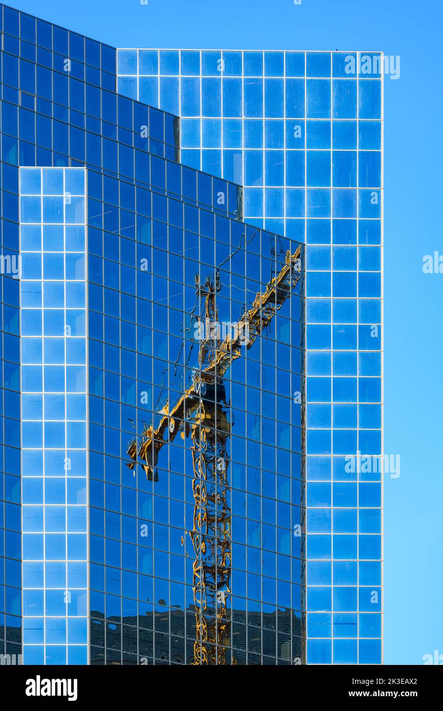 Tall yellow crane reflecting on a skyscraper with glass windows made blue by the clear summer sky Stock Photo