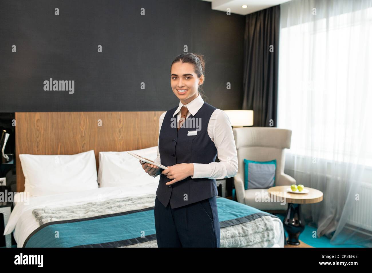 Photo realistic digital collage of happy young female worker of luxurious hotel in uniform standing in front of camera against bed Stock Photo