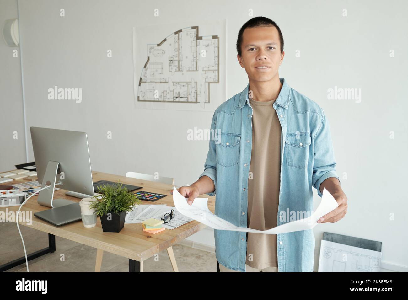 Photo realistic digital collage of young Chinese creative designer with blueprint standing against his workplace Stock Photo