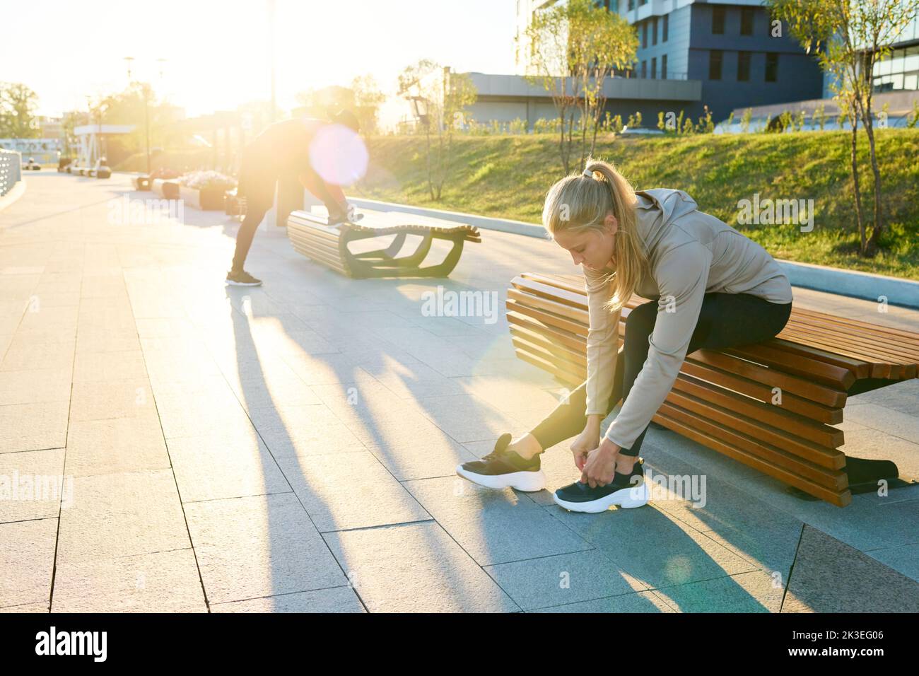 Young blond sportswoman in activewear sitting on bench in urban environment and bending over sneaker while tying shoelace Stock Photo